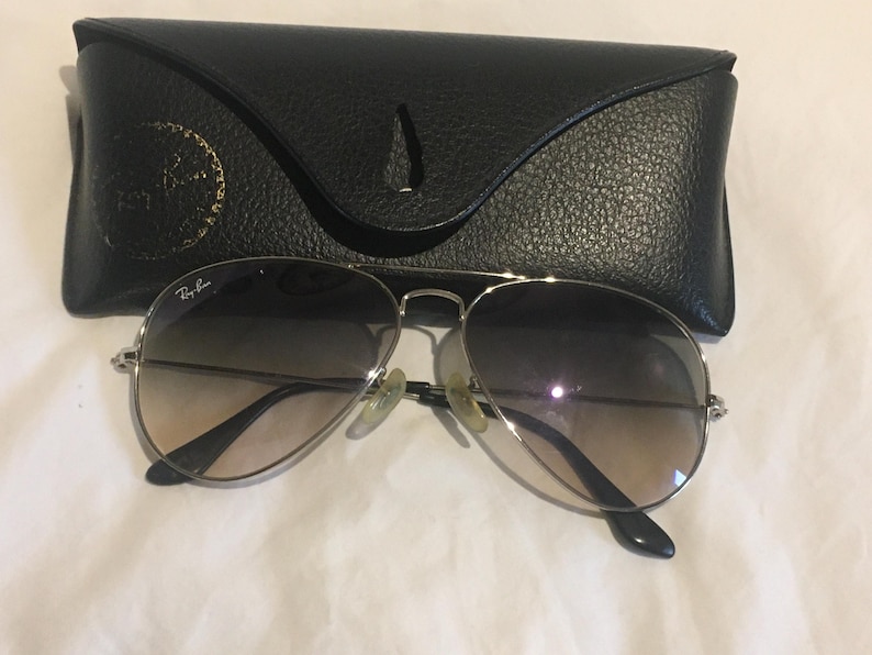 K&ouml;nnte beinhalten: Silberne Aviator-Sonnenbrille mit braunen Verlaufsgl&auml;sern und einem schwarzen Lederetui. Das Etui tr&auml;gt das Ray-Ban-Logo auf der Vorderseite.