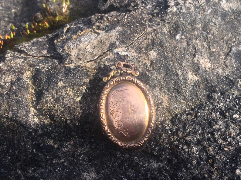 May include: An oval-shaped gold locket with an ornate design and a floral pattern engraved on the front.