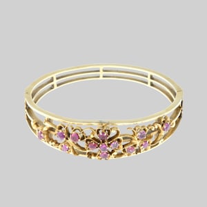 May include: A gold-coloured hinged bangle bracelet with a floral design. The bracelet features pink gemstones set within the floral pattern. The bracelet has a three-bar design on the top.