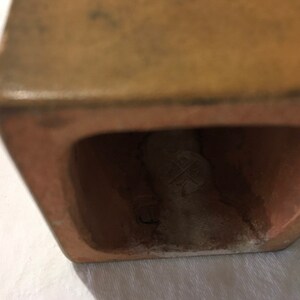 Op de afbeelding: Close-up van de onderkant van een terracottakleurige keramische plantenbak. De plantenbak heeft een vierkante vorm en een kleine opening aan de onderkant. De opening onthult een wit keramisch merkteken met een kruis erin.