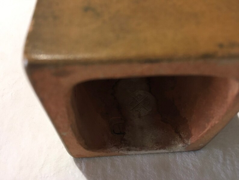 Op de afbeelding: Close-up van de onderkant van een terracottakleurige keramische plantenbak. De plantenbak heeft een vierkante vorm en een kleine opening aan de onderkant. De opening onthult een wit keramisch merkteken met een kruis erin.