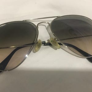 K&ouml;nnte beinhalten: Silberne Aviator-Sonnenbrille mit braunen Verlaufsgl&auml;sern. Die Sonnenbrille hat ein Ray-Ban-Logo auf der linken Linse.