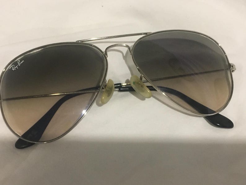 K&ouml;nnte beinhalten: Silberne Aviator-Sonnenbrille mit braunen Verlaufsgl&auml;sern. Die Sonnenbrille hat ein Ray-Ban-Logo auf der linken Linse.