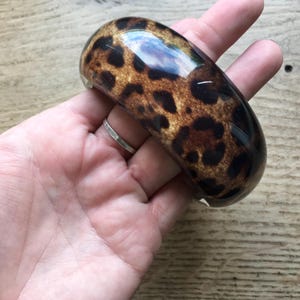 May include: A glossy, wide bangle bracelet with a leopard print pattern in shades of brown and black. The bracelet is held in a hand, showcasing its smooth, curved design. This statement piece is a bold accessory.