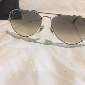 K&ouml;nnte beinhalten: Silberne Aviator-Sonnenbrille mit braunen Verlaufsgl&auml;sern. Die Sonnenbrille liegt auf einer wei&szlig;en Oberfl&auml;che.