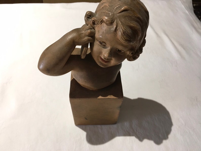 Op de afbeelding: Een terracotta buste van een jong meisje met kort krullend haar, haar hand aan haar oor houdend, op een vierkante basis.