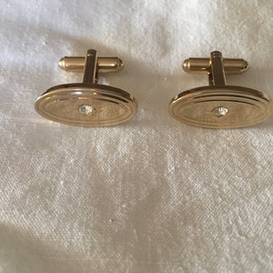 Puede incluir: Un par de gemelos ovalados de oro con un solo strass transparente en el centro de cada gemelo.