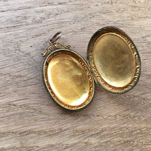 May include: A gold-toned oval locket with an ornate design around the edges. The locket is open, revealing a blank interior.