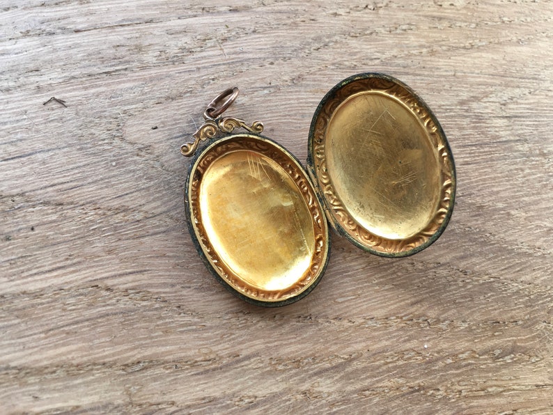 May include: A gold-toned oval locket with an ornate design around the edges. The locket is open, revealing a blank interior.