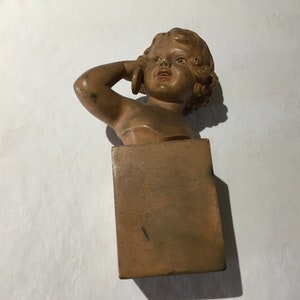 Op de afbeelding: Een terracotta buste van een jong meisje met krullend haar, dat een eenvoudige jurk draagt. De buste is gemonteerd op een vierkante houten voet.