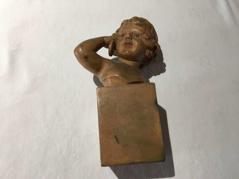 Op de afbeelding: Een terracotta buste van een jong meisje met krullend haar, dat een eenvoudige jurk draagt. De buste is gemonteerd op een vierkante houten voet.