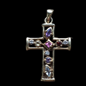 Peut inclure: Pendentif en forme de croix de couleur or, orné de diverses pierres précieuses. Les pierres précieuses comprennent des pierres violettes, roses, rouges et transparentes. La croix est posée sur un fond noir.