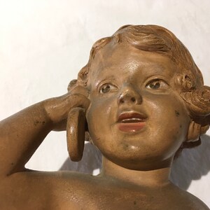 Op de afbeelding: Een terracotta beeld van een cherubijn met een ring in zijn hand, de cherubijn heeft een verraste uitdrukking op zijn gezicht.