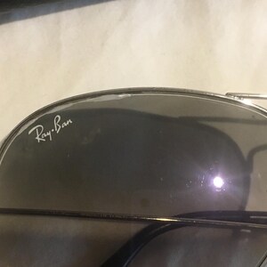 K&ouml;nnte beinhalten: Silberne Aviator-Sonnenbrille mit dunkelgrauen Gl&auml;sern. Die Sonnenbrille tr&auml;gt den Schriftzug "Ray-Ban" auf der linken Linse.