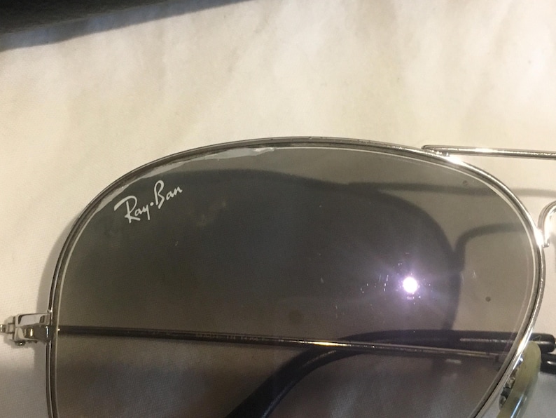K&ouml;nnte beinhalten: Silberne Aviator-Sonnenbrille mit dunkelgrauen Gl&auml;sern. Die Sonnenbrille tr&auml;gt den Schriftzug "Ray-Ban" auf der linken Linse.