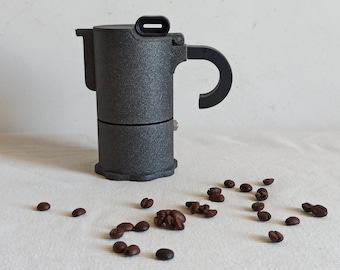 May include: A gray metal stovetop espresso maker with a black handle and spout. Coffee beans are scattered around the maker on a white surface.