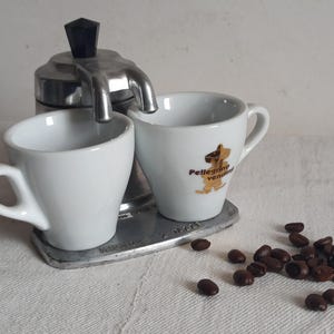 Puede incluir: Una pequeña cafetera espresso plateada con dos tazas de cerámica blancas. Una taza tiene un logotipo con texto y una ilustración de perro. Granos de café esparcidos junto a las tazas. La cafetera tiene un asa negra.