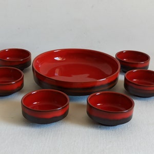 Può includere: Un set di sette ciotole in ceramica. Il set comprende una grande ciotola poco profonda e sei ciotole più piccole. Tutte le ciotole hanno un interno rosso lucido e una fascia marrone scuro alla base.