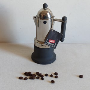 May include: A silver and black stovetop espresso maker with a black handle and a black base. The maker has a label that reads "vev VIGANO".