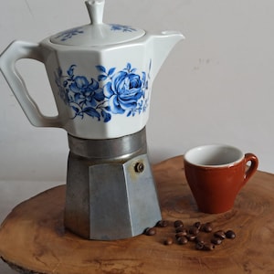 Puede incluir: Una cafetera italiana vintage con una tapa de porcelana blanca con diseños florales azules. Una pequeña taza de cerámica marrón y granos de café sobre una superficie de madera.