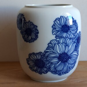 May include: A white ceramic vase with a rounded form, decorated with a blue floral design. The flowers are depicted with intricate line art. The vase has a glossy finish and is suitable for displaying flowers or as a decorative item.