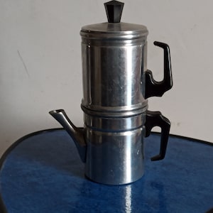 May include: A silver and black stovetop espresso maker with a black handle and spout. The maker is sitting on a blue surface.
