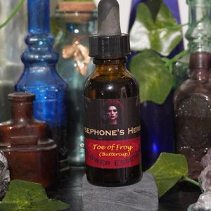May include: A brown glass bottle with a dropper top and a black label that reads "Sephone's Herbs Toe of Frog (Buttercup) Flower Essence". The bottle is surrounded by various glass bottles and crystals.