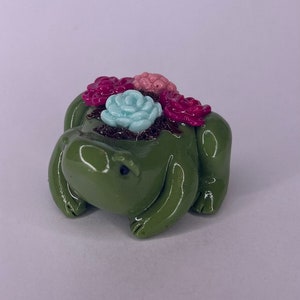 May include: A green ceramic frog figurine with pink, red, and blue flowers on its head. The frog is sitting on its belly with its legs tucked underneath it.