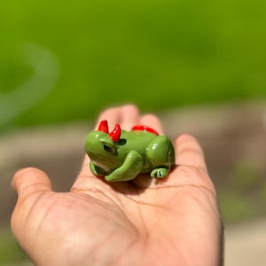 May include: A green frog figurine with red horns and a red spine. The frog is in a sitting position with its front legs bent. The figurine is small and appears to be made of ceramic or a similar material.