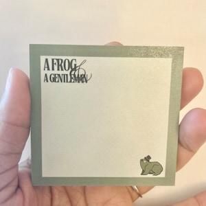 May include: Square, sage green notepad with the text "A Frog & A Gentleman" in black. A small illustration of a frog wearing a top hat is in the bottom right corner. The notepad is held in a hand.