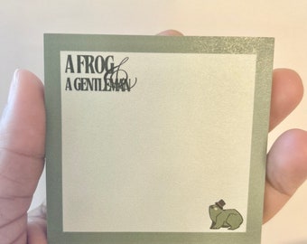 May include: Square, sage green notepad with the text "A Frog & A Gentleman" in black. A small illustration of a frog wearing a top hat is in the bottom right corner. The notepad is held in a hand.