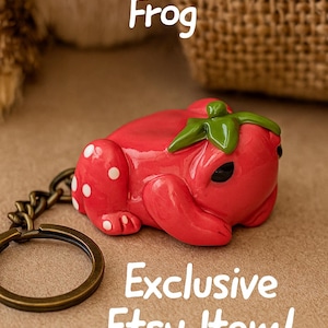 May include: A red strawberry frog keychain with white polka dots and green leaves. The text "Strawberry Frog" and "Exclusive Etsy Item!" are displayed on the image. The keychain has a bronze-colored ring and chain.