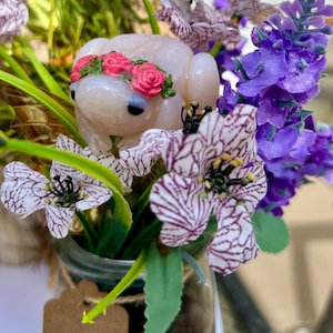 以下が含まれることがあります： ピンクの花の冠をかぶった小さな白いガラスの蛙の置物が、紫と白の花が入ったガラス瓶に入っています。