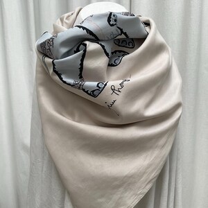 Op de afbeelding: Een beige zijden sjaal met een lichtblauw patroon. De sjaal is gedrapeerd en toont een zachte, vloeiende textuur. Het lichtblauwe gedeelte heeft een zwarte illustratie en de handgeschreven handtekening "Juin Phony".