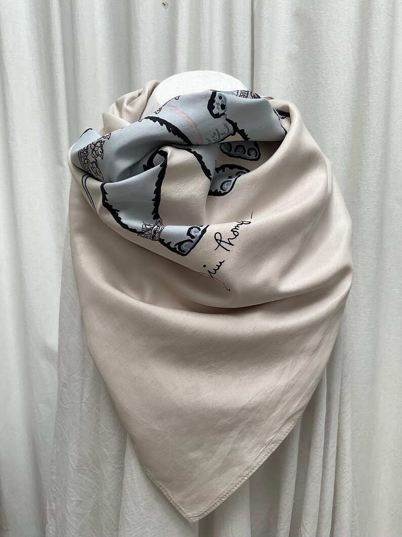 Op de afbeelding: Een beige zijden sjaal met een lichtblauw patroon. De sjaal is gedrapeerd en toont een zachte, vloeiende textuur. Het lichtblauwe gedeelte heeft een zwarte illustratie en de handgeschreven handtekening "Juin Phony".