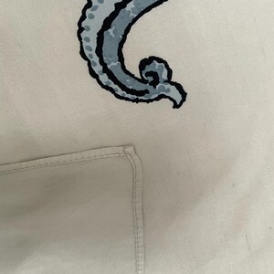 Op de afbeelding: Lichtbeige textiel met een groot, gestileerd, gebogen ontwerp in blauw- en zwarttinten. Het ontwerp heeft een gestippeld patroon en een wervelend uiteinde. Een gevouwen hoek van het textiel is linksonder zichtbaar.