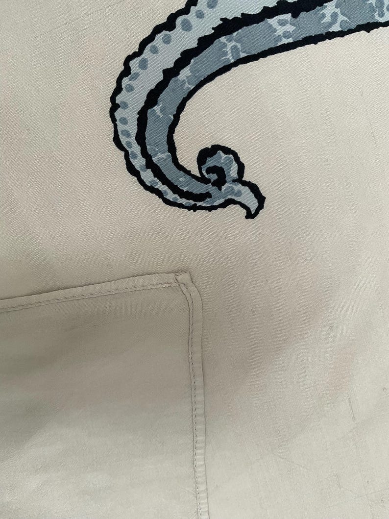 Op de afbeelding: Lichtbeige textiel met een groot, gestileerd, gebogen ontwerp in blauw- en zwarttinten. Het ontwerp heeft een gestippeld patroon en een wervelend uiteinde. Een gevouwen hoek van het textiel is linksonder zichtbaar.