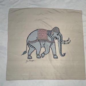 Op de afbeelding: Een beige vierkant kussen met een gedetailleerde illustratie van een grijze olifant, versierd met een roze en grijs patroon zadel en decoratieve hoofddeksels. De olifant heeft grote slagtanden en loopt naar links. De handtekening van de kunstenaar staat onderaan.