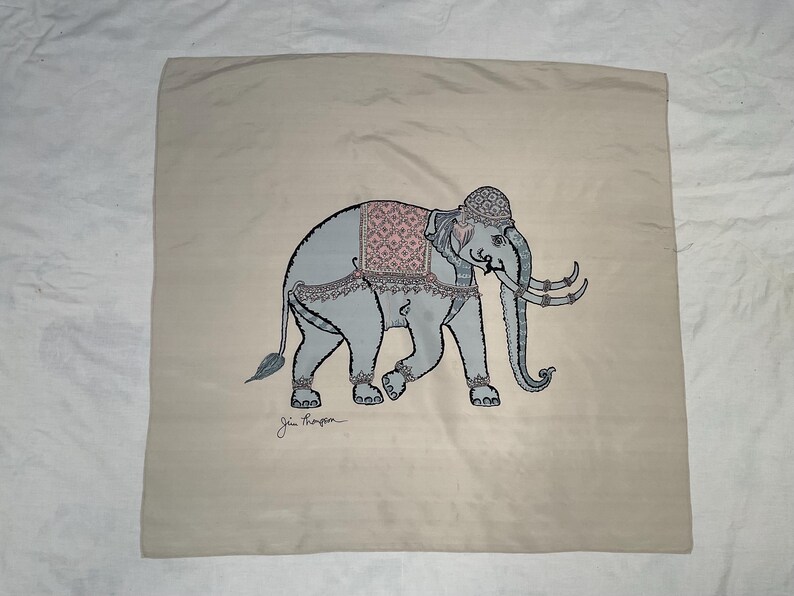 Op de afbeelding: Een beige vierkant kussen met een gedetailleerde illustratie van een grijze olifant, versierd met een roze en grijs patroon zadel en decoratieve hoofddeksels. De olifant heeft grote slagtanden en loopt naar links. De handtekening van de kunstenaar staat onderaan.