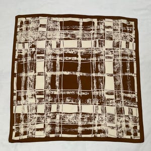 May include: A square scarf in brown and cream, featuring a grid design. The scarf has a dark brown border and a repeating pattern of cream lines on a brown backdrop. The design is abstract and textured.