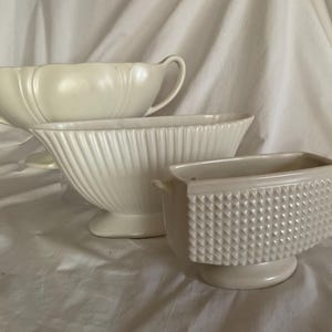 May include: Three cream-coloured ceramic planters of varying sizes. The largest has handles and a scalloped rim. The middle planter has vertical ridges, and the smallest is rectangular with a textured, diamond pattern.