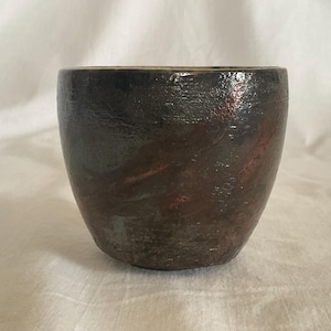 May include: A handmade ceramic cup with a dark, textured exterior. The cup has a rounded shape and a dark brown color with hints of red and green. The interior is a lighter brown. The cup is suitable for beverages.