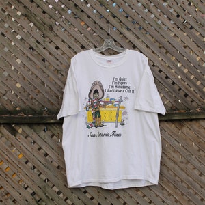 May include: White t-shirt with a cartoon illustration of a man wearing a sombrero and holding a bottle of beer. The text on the shirt reads "I'm Quiet I'm Happy I'm Handsome I don't give a Chit!!" and "San Antonio, Texas".