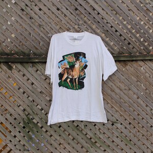 Pode incluir: Camiseta branca com uma estampa gr&aacute;fica de um veado com chifres em um ambiente de floresta. O veado &eacute; cercado por veados menores no fundo.