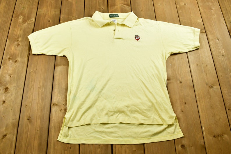 K&ouml;nnte beinhalten: Ein gelbes Poloshirt mit kurzen &Auml;rmeln und einem gestickten roten und wei&szlig;en Comic-Charakter auf der linken Brust. Das Shirt hat einen gr&uuml;nen Kragen und einen Knopfverschluss.