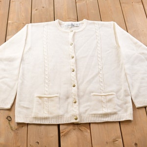 Könnte beinhalten: Ein weißer Strickcardigan mit Zopfmuster und goldenen Knöpfen. Der Cardigan hat zwei Taschen auf der Vorderseite.