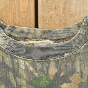 May include: Close-up of a camouflage-patterned sweatshirt collar. The collar is ribbed and gray, with a faded camouflage design in shades of green, brown, and tan. The fabric appears worn, suggesting a vintage or pre-owned item.