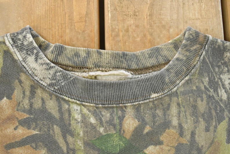 May include: Close-up of a camouflage-patterned sweatshirt collar. The collar is ribbed and gray, with a faded camouflage design in shades of green, brown, and tan. The fabric appears worn, suggesting a vintage or pre-owned item.
