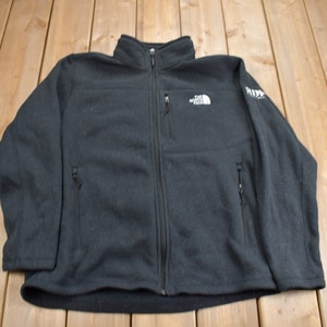 Könnte beinhalten: Schwarze Fleecejacke mit durchgehendem Reißverschluss, zwei Seitentaschen und einer Brusttasche. Das The North Face-Logo ist auf der Brust sichtbar. Die Jacke hat einen hohen Kragen und lange Ärmel.