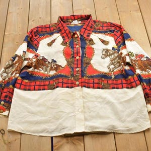 May include: A long-sleeved button-down shirt with a white base and a red, blue, and white plaid pattern. The shirt features a repeating pattern of horses and riders, with a gold chain design.